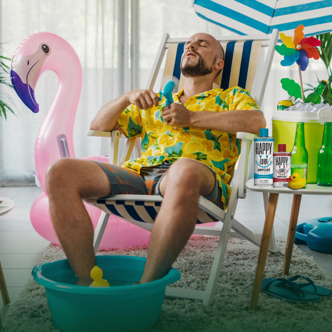 A relaxed man in tropical clothes lounges in a deck chair indoors with his feet in a small inflatable pool, inflatable flamingo beside him, and Happy Lube bottles on a table — capturing a playful staycation vibe with fun, flirty summer energy.