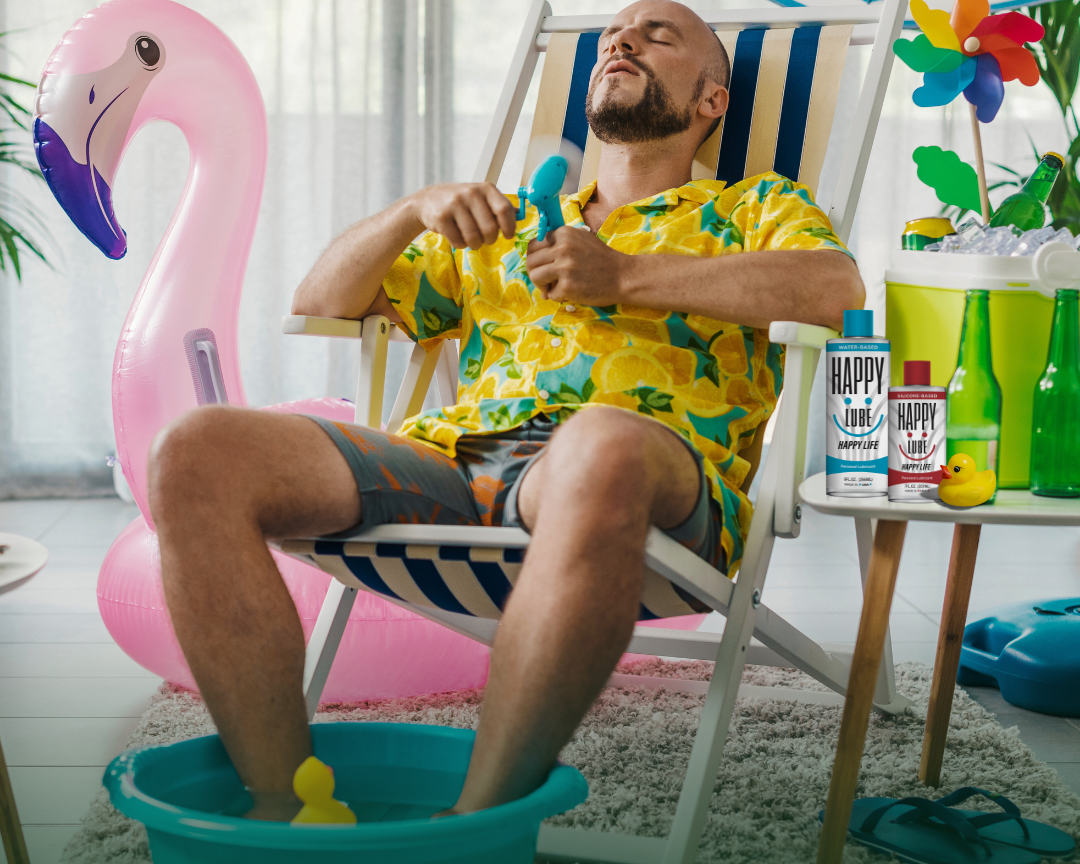 A relaxed man in tropical clothes lounges in a deck chair indoors with his feet in a small inflatable pool, inflatable flamingo beside him, and Happy Lube bottles on a table — capturing a playful staycation vibe with fun, flirty summer energy.